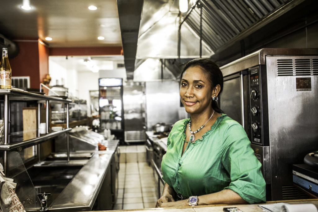 An African American woman with a emerald colored blouse and matching necklace stands in a restaurant kitchen.