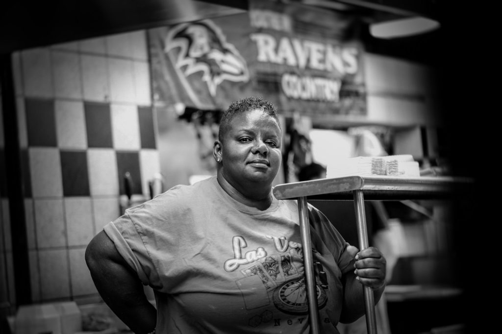 An African American woman with a Las Vegas t-shirt stands in a restaurant with a Ravens Country banner on the wall.