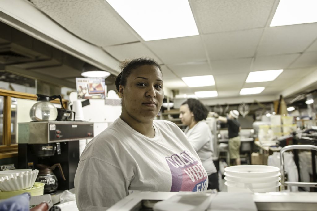 An African American woman stands next to a coffee maker in a kitchen environment.