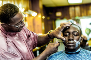 An African American man with short hair and glasses shaves a man in barber shop.