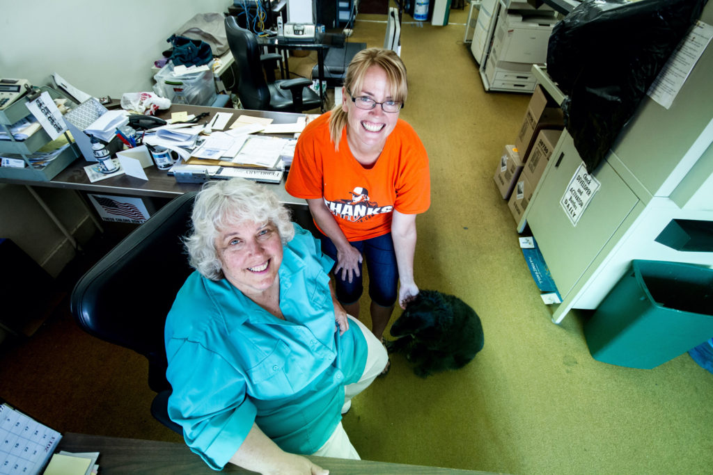 Two white women, one wearing a teal-blue shirt and one with a bright orange shirt, look up at the camera in their printing office.