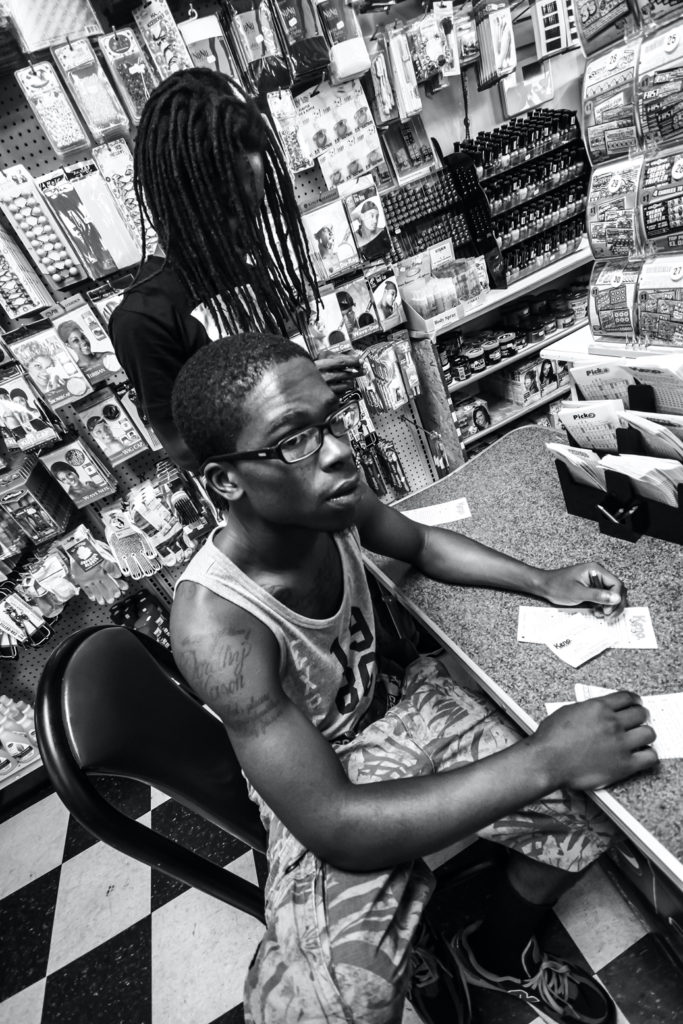 An African American young man with a tank top and black glasses sits at a desk in a market, surrounded by products.