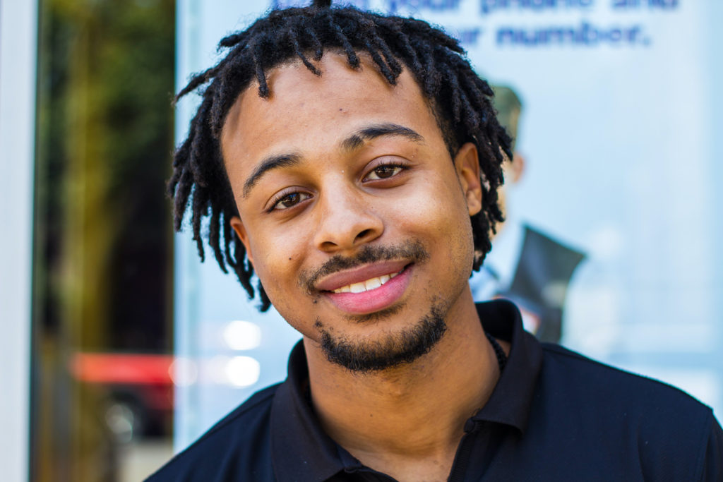 An African American man with short dreadlocks and a goatee wears a black collared shirt and smiles at the camera.