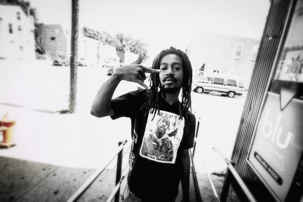 An African American man with dreadlocks, a beard, and a black t-shirt makes a sign with his thumb, index finger and little finger.