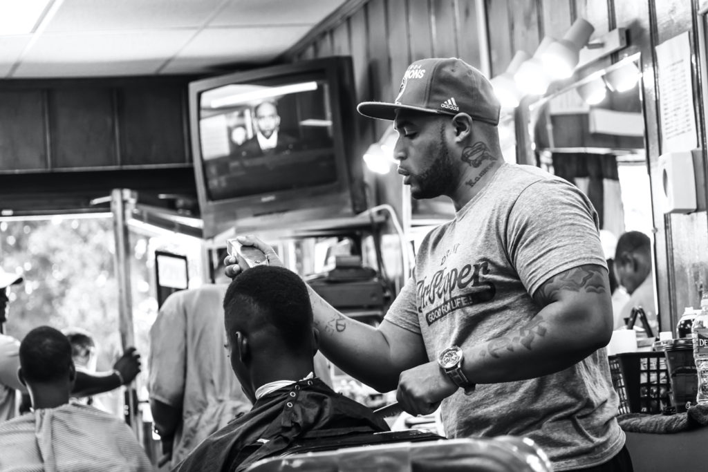 An African American man, wearing a ball cap and a gray t-shirt, works with a client in a barber shop.
