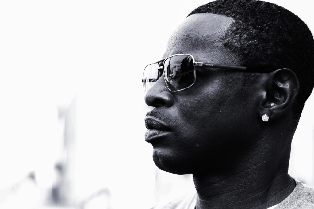 An African American man looks away from the camera as he sports a stylish pair of sun glasses and diamond stud earring.