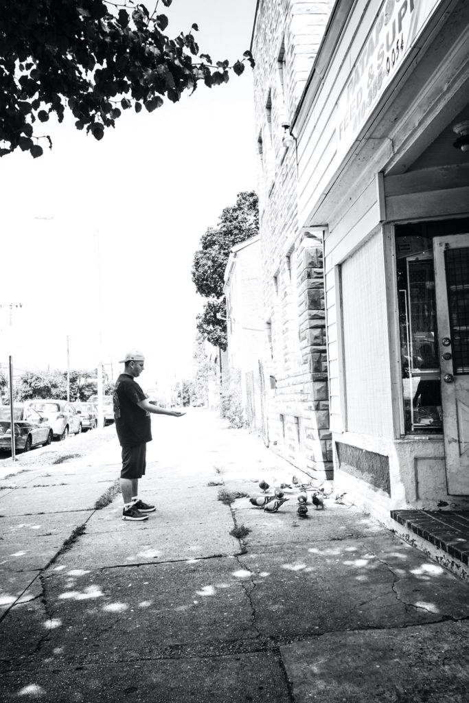 A white man, wearing a backwards baseball cap, stands on the sidewalk and throws seeds to a number of pigeons.