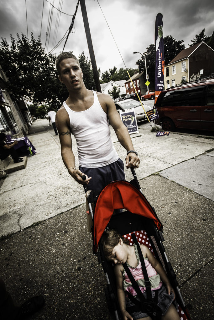 A white young man in a white tank top pushes a little girl riding in a red stroller.