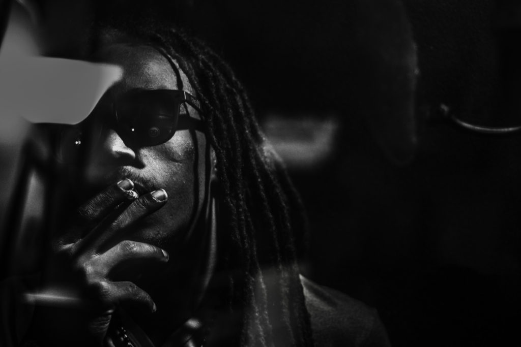 An African American man with long dreadlocks and black glasses smokes a cigarette.