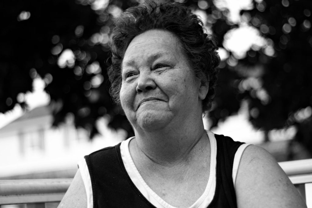 An older white woman, wearing a black-and-white tank top, smiles as she sits under the canopy of a tree.