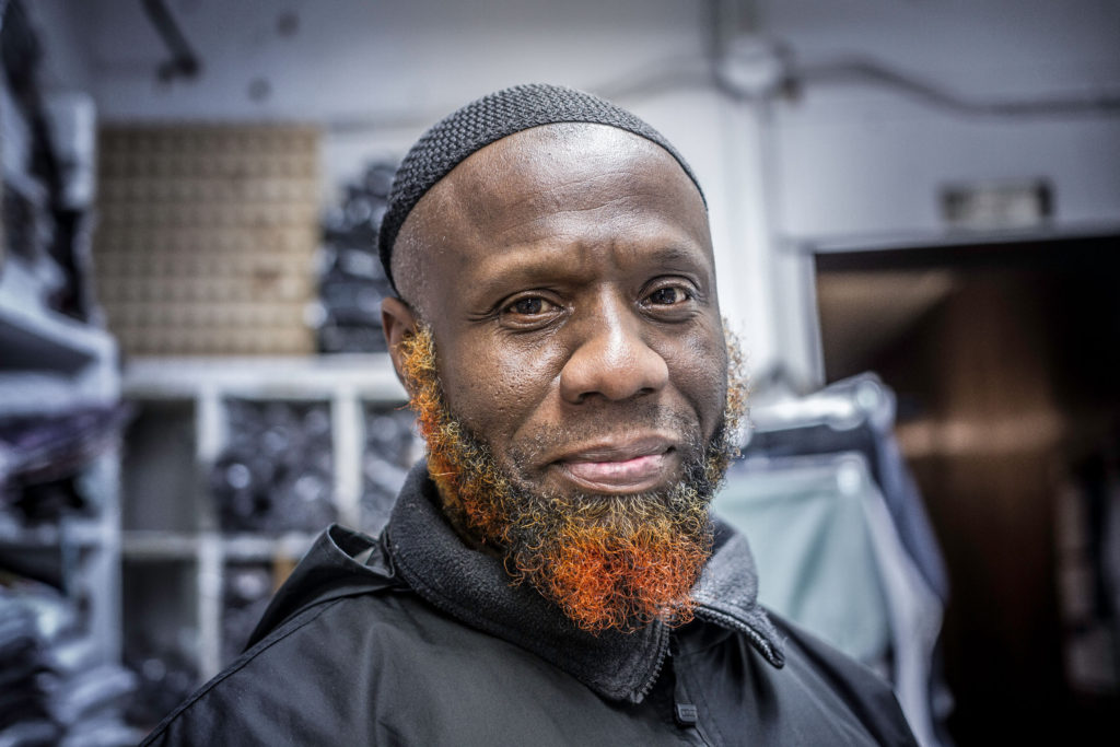 An African American man with a tight black cap and a colorful orange and black beard smiles at the camera.