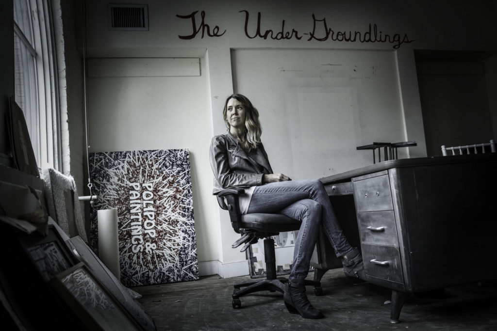 A white woman with jeans and a gray blazer sits in an office chair, surrounded by artwork in a rundown office space.