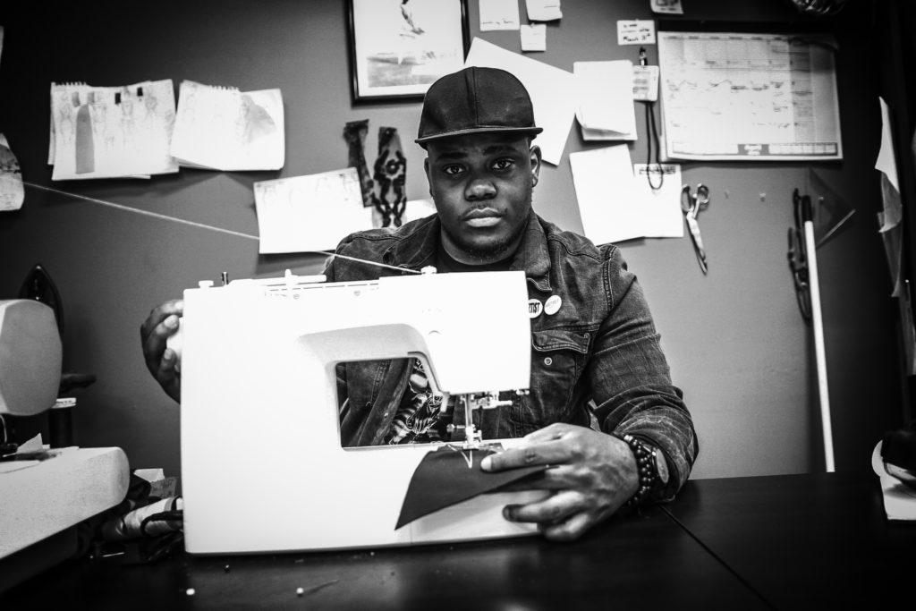 An African American man wearing a jeans jacket and black ballcap uses a white sewing machine.
