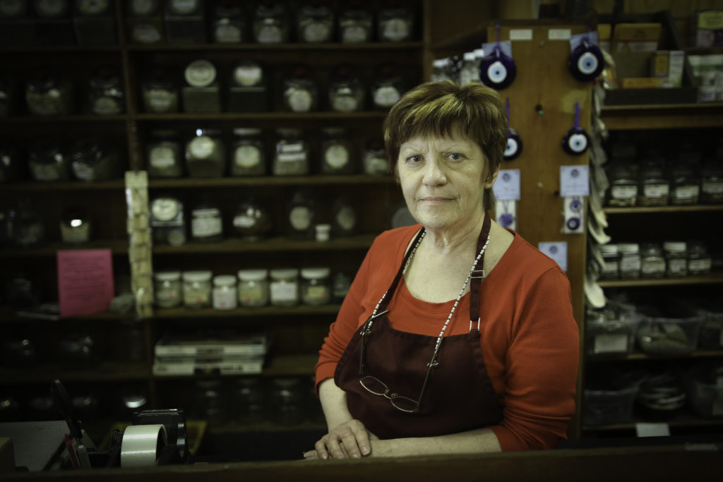 An older white woman with a red shirt and apron stands behind the counter of shop with candles and herbs on the shelves.