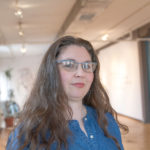 A white woman wearing artsy glasses and denim shirt stands in what appears to be a gallery space.