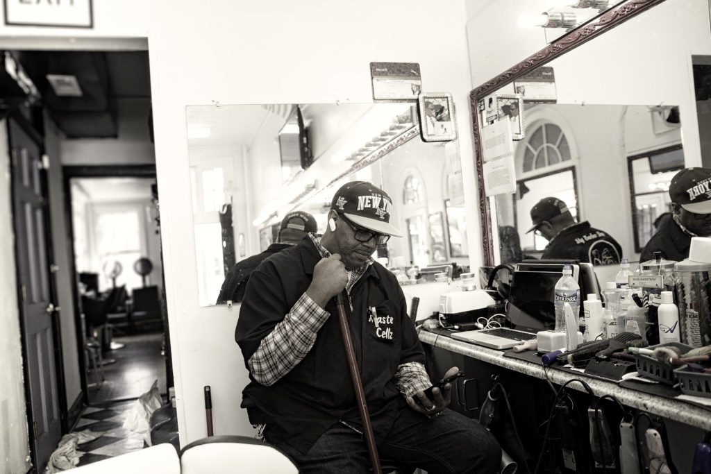 An African American man with a New York, New York hat checks his phone as he sits in a chair at a barbershop.