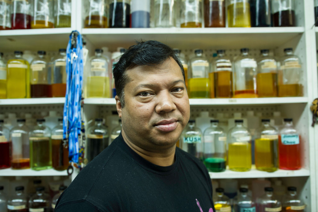 A man with a black t-shirt stands in a pharmacy or other location with color bottles of liquids behind him.