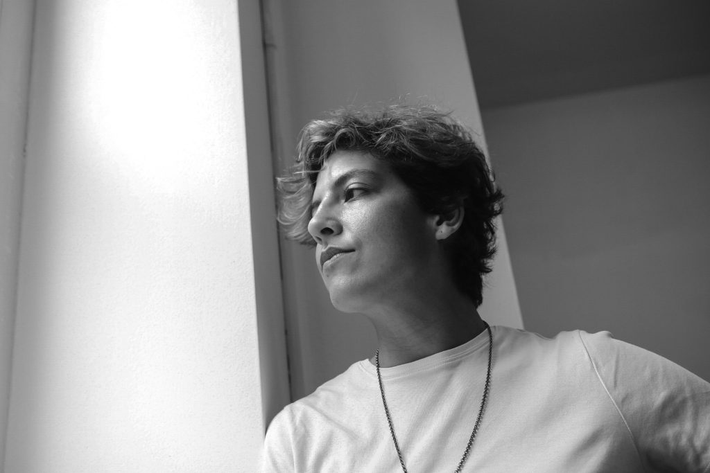 A woman with tussled, light brown hair and a white t-shirt looks pensively out of the window.
