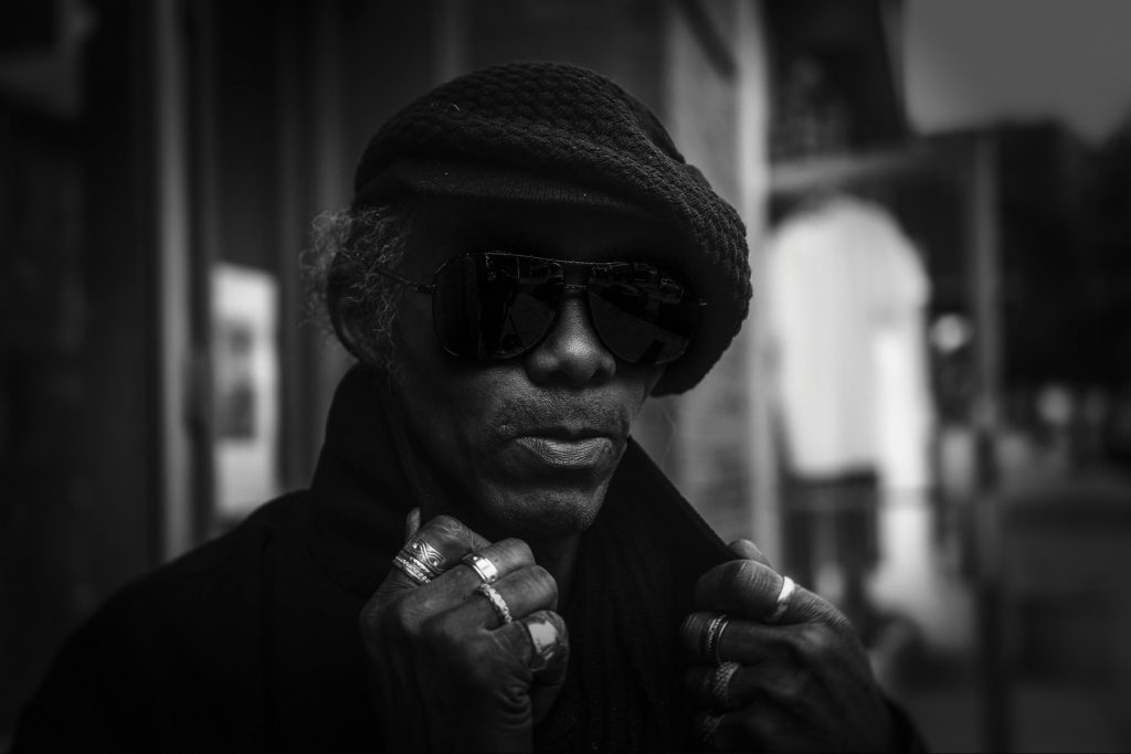 An older African American man with a black beret and sunglasses holds the collar of a his shirt, revealing a ring on every finger of his hands.