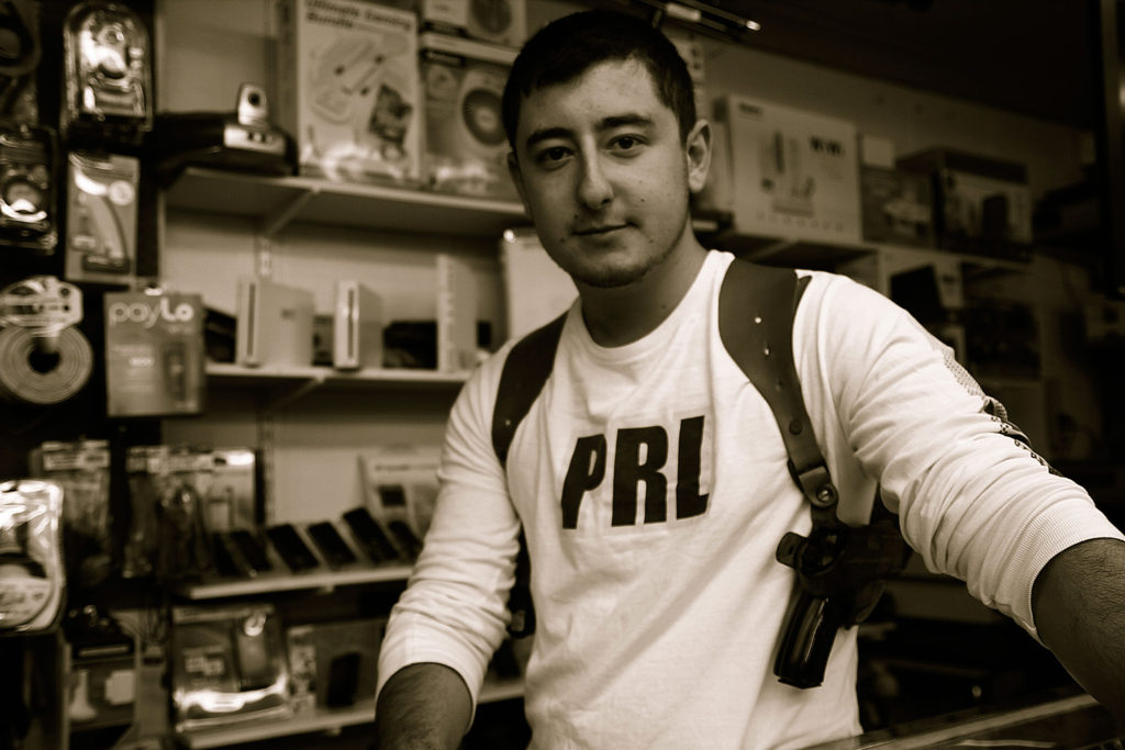 A white man with white t-shirt that says PRL stands behind a counter as he wears a holster with a handgun.