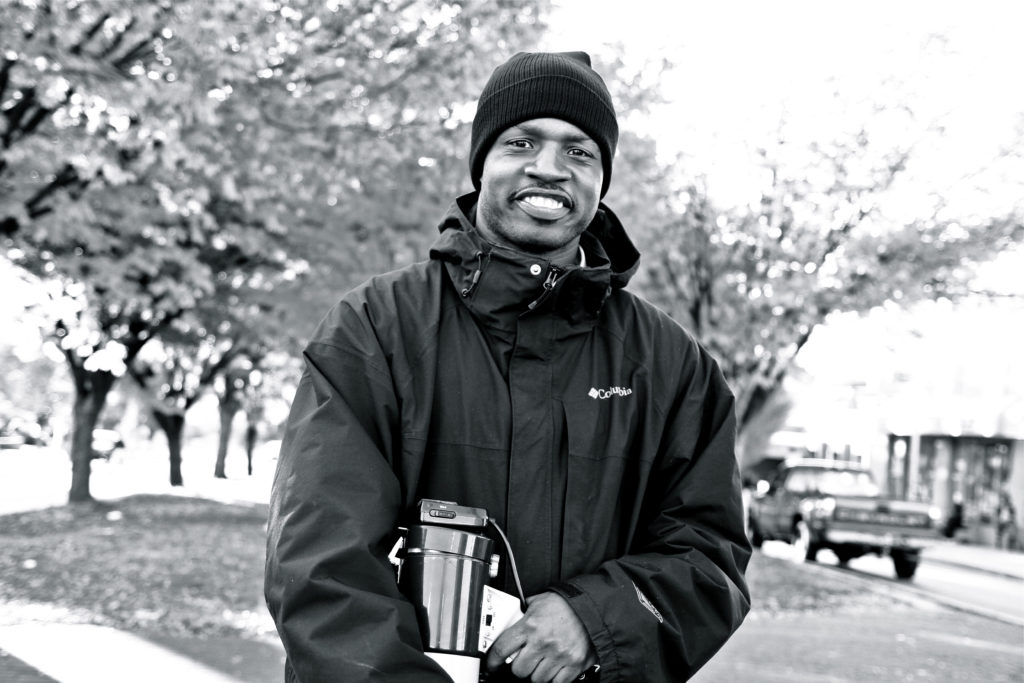 An African American man wears a black jacket and knit cap as he holds a metal object like a paint canister or sprayer.