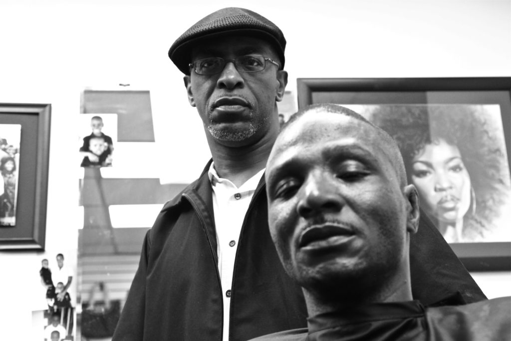Two African American men look down at the camera. One appears to be getting a haircut while the other looks on.