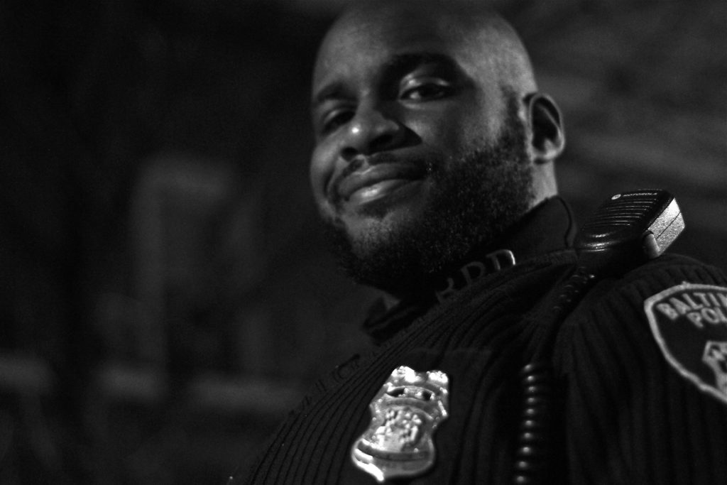 An African American male police officer smiles as he displays his badge and a radio on his shoulder.