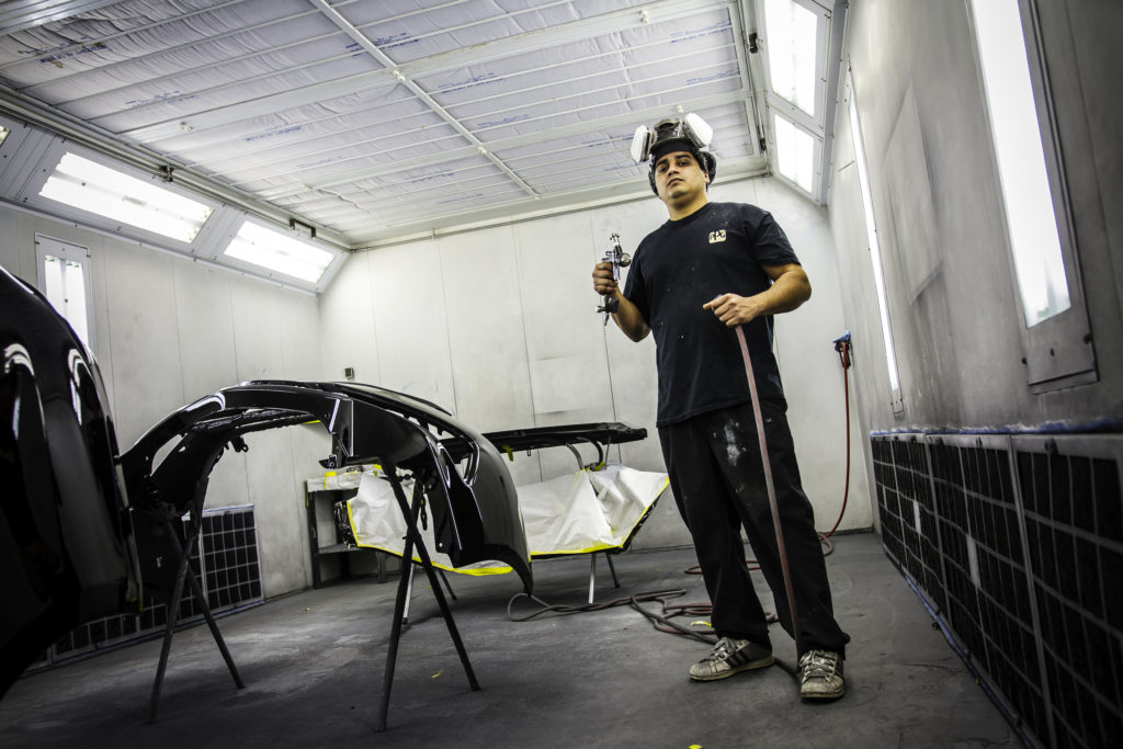 A man dressed in black holds a paint sprayer in an auto body shop.