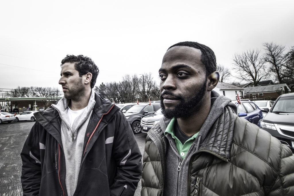 A white man on the left in a gray jacket and an African American man on the right in a brown puffy coat, stand in a car parking lot.