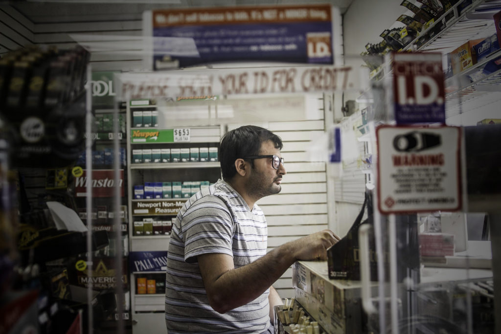A white man with Buddy Holly glasses and a striped shirt sits at the cash register in a market.