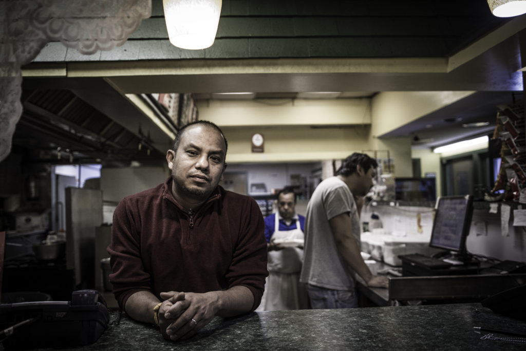 A man with a dark red pullover stands at a counter in a restaurant kitchen and looks out at the camera.
