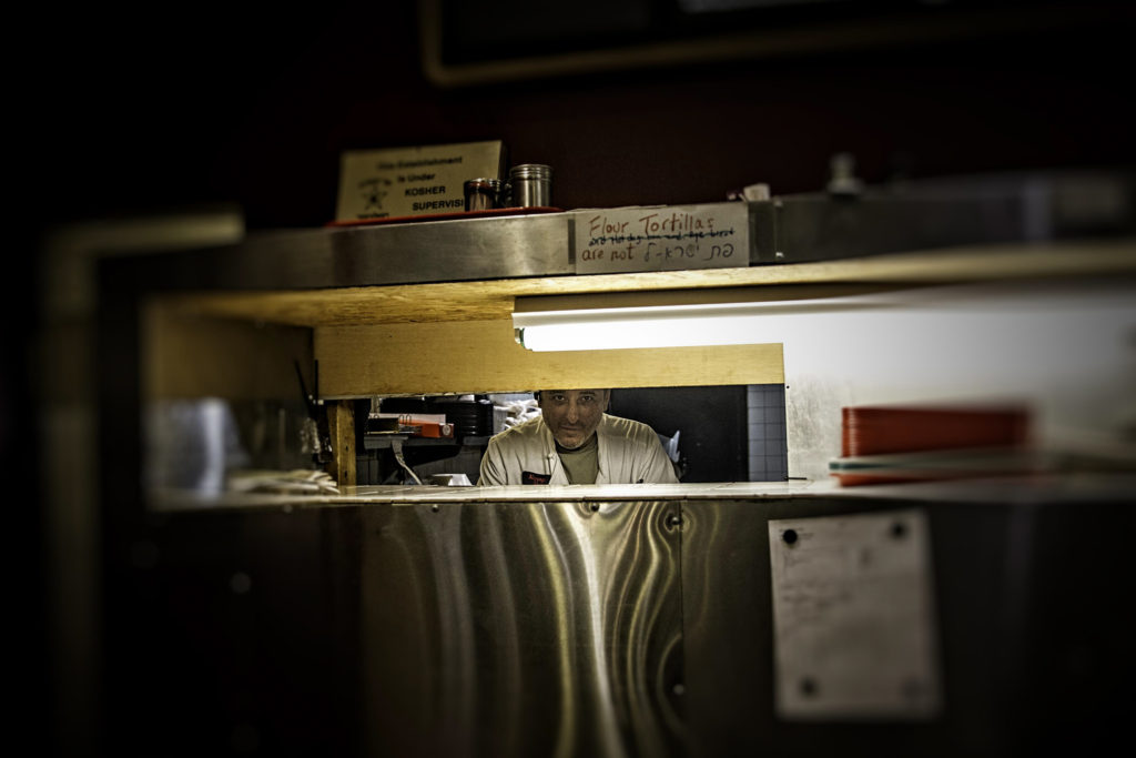A white man in a white jacket looks through a narrow window at a restaurant kitchen.