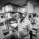 An African American man with a striped golf shirt and a ballcap cleans the grill in a kitchen.