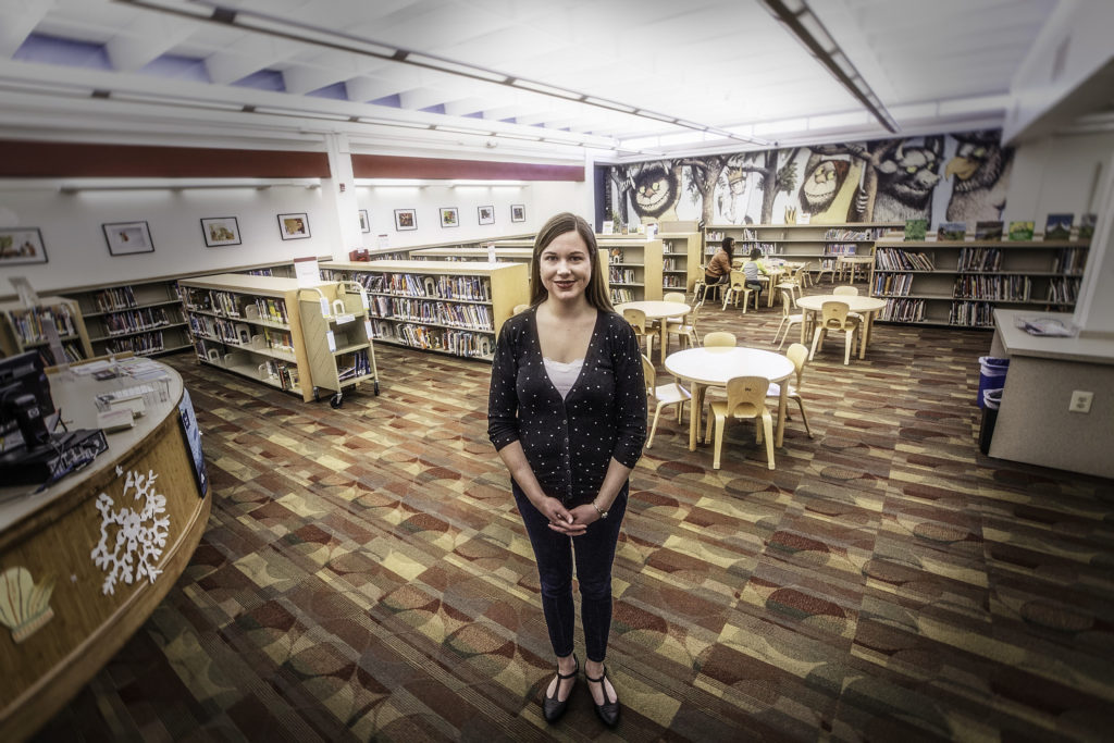 A white woman with a black polka dotted shirt stands in a library with a colorfully carpeted floor.
