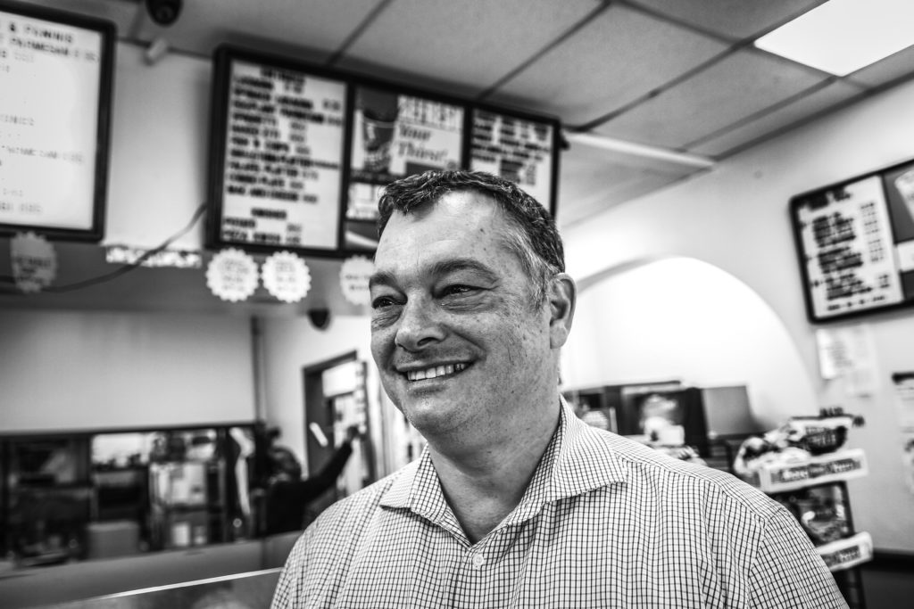A white man with a collared checked shirt stands at the counter of a takeout restaurant and smiles.