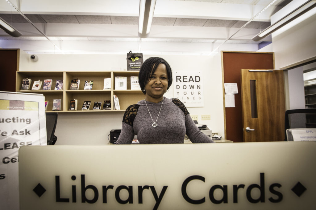 An African American woman with a gray shirt and silver necklace stands behind a library counter.