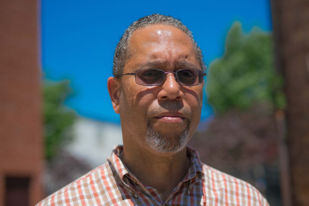 An African American man with a plaid, button-down shirt and sun glasses looks at the camera. 