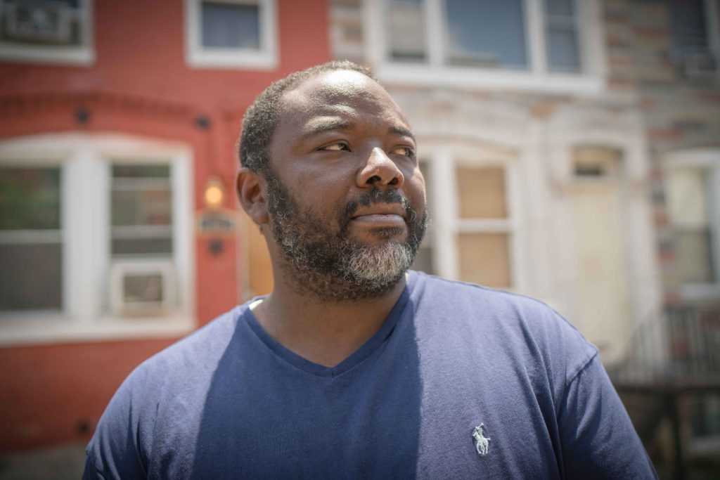 An African American man with a blue t-shirt stands in front of row houses as he looks off to the side. 