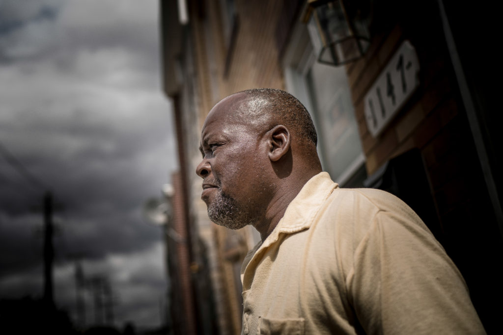 An African American man with a bald head and a beige, collared shirt leans against a brick building.