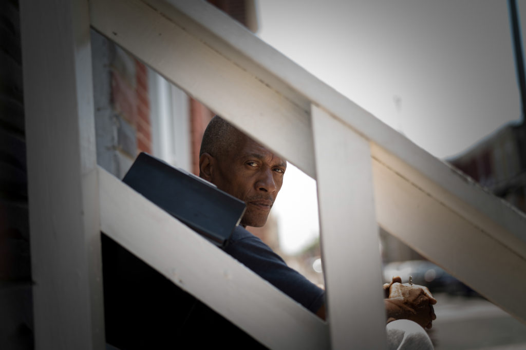 An African American man looks through the slats of a railing as he sits on the steps of a building. 