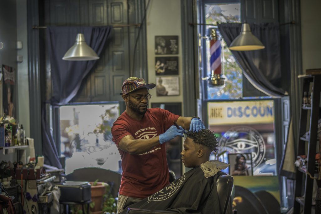 An African American man with a bright, red t-shirt and brown ballcap cuts the hair of another young man in a barber shop.