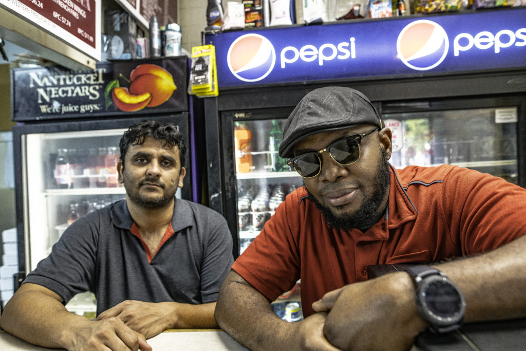 Two men, one with a gray collared shirt and the other with a gray cap and dark sunglasses, stand in a small market with a Pepsi sign behind them.