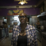 An African American woman with Buddy Holly-style glasses and a plaid shirt stands in a restaurant kitchen.