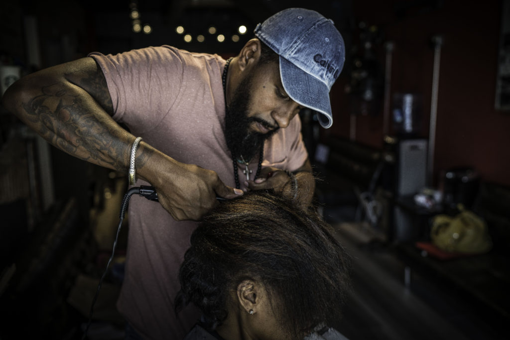 A man with a dark beard and blue ballcap cuts a woman's hair.