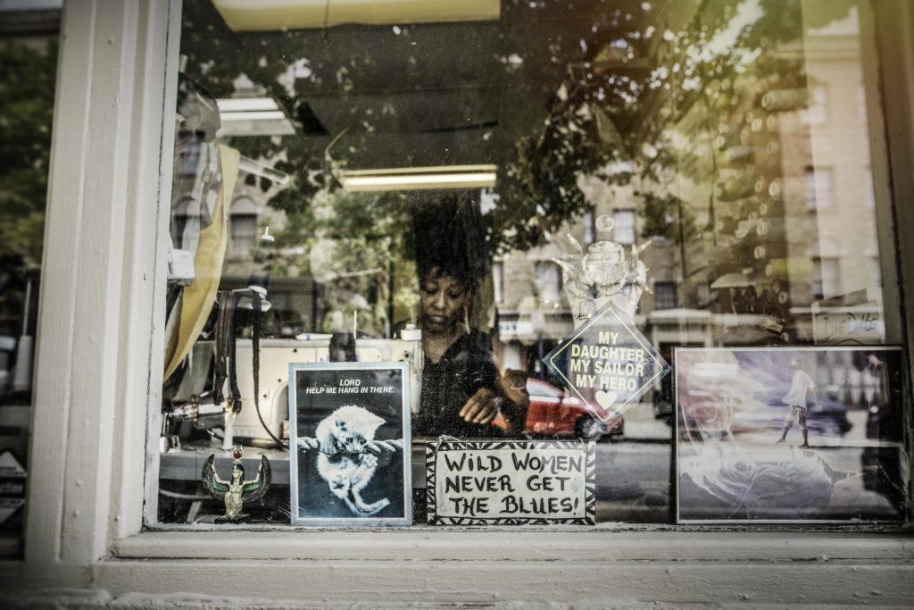 A woman with black hair appears behind the glass of a shop window with various signs in it, one reading, Wild Women Never Get the Blues.