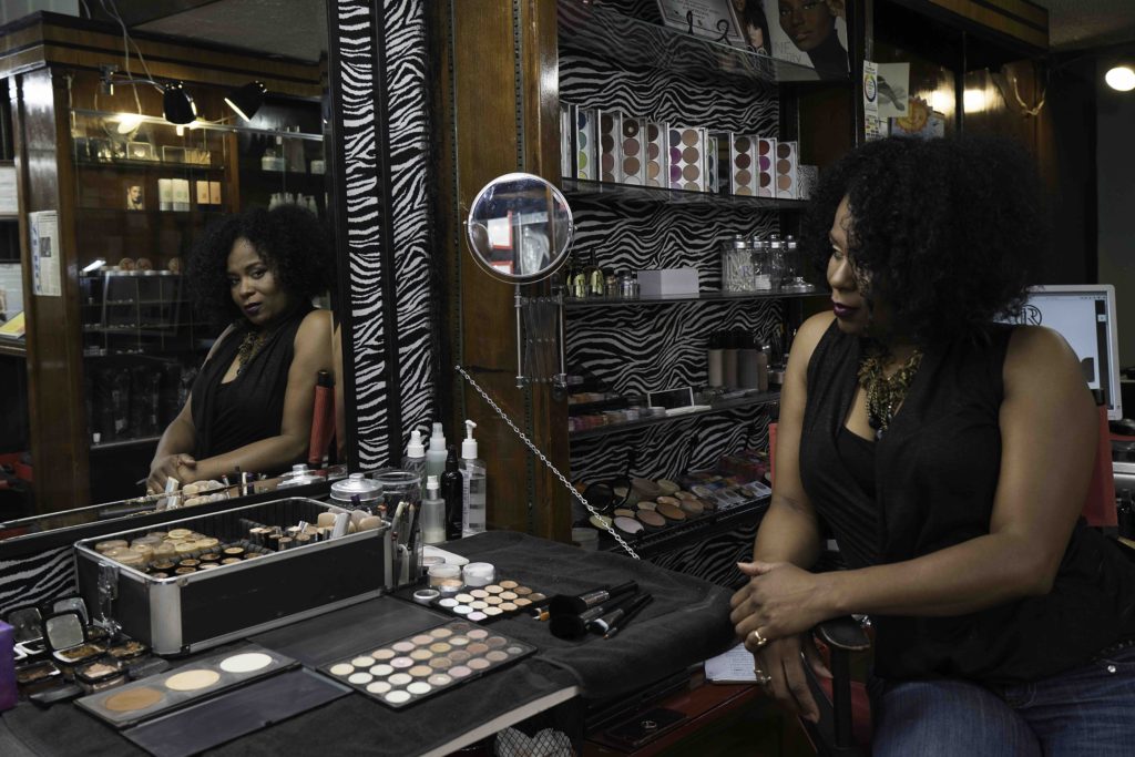 A woman with black curly hair stands in front of a station in a nail salon with make up and hair products.