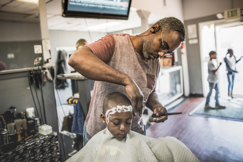 An African American man with glasses cuts a little boy's hair in a barber shop.
