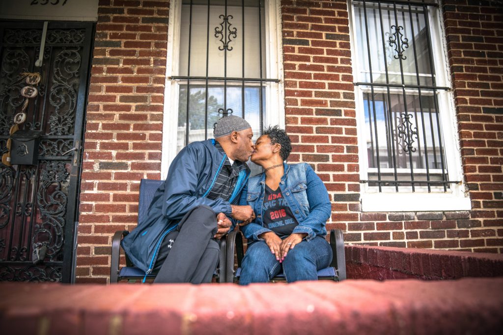 Two people lean in for a kiss as they sit on the front porch of a brick house.