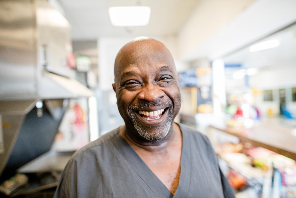 An African American man with a gray v-neck shirt smiles broadly inside a restaurant setting.