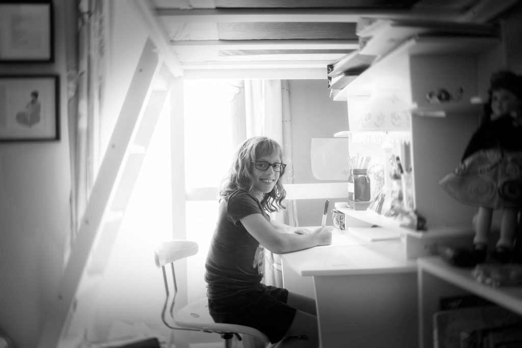 A young girl with light hair and glasses sits at a desk in